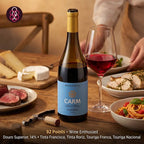 A bottle of Carm Reserva Douro red wine stands on a wooden dining table beside a filled wine glass and plated food, with a person partially visible in the background. The bottle label reads 'Reserva Carm' and 'Douro 2023'.