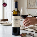 A bottle of 2022 Vertente Douro Non-Alcoholic Red Wine from Serrapí, Portugal, standing on a marble countertop next to a hand holding a glass of the deep red wine. A corkscrew is in the foreground, and a blurred kitchen background is visible.