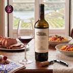 A bottle of Quinta Da Romaneira Dona Clara red wine with a glass of red wine, a corkscrew, and bowls of pasta and sliced meat on a table in front of a window with a vineyard view. A logo for 'O'N' appears in the top left corner.