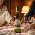 A bottle of S
Calem Velhotes white wine is displayed on a bed with two filled wine glasses, a cork, and a woman and man partially visible in the background, creating a cozy, warm atmosphere.