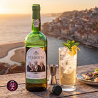 A bottle of Calém Velhotes white wine stands on a wooden table beside a glass of iced cocktail with mint and a lemon twist, with a jigger and cork. The background shows a coastal city at sunset with the sea and shoreline visible.