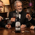 A bottle of Calem Velhotes red wine is displayed on a wooden table, surrounded by three men in black suits holding wine glasses and one pouring wine. The wine bottle has a white label with black text and a portrait of three men.