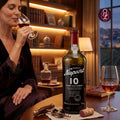 A bottle of Porto Nepoort 10 Years Old Tawny Port wine is displayed on a table in a warm, elegant room setting, with a woman in a dark robe holding a glass of red wine in the background. Two corks are placed in the foreground.