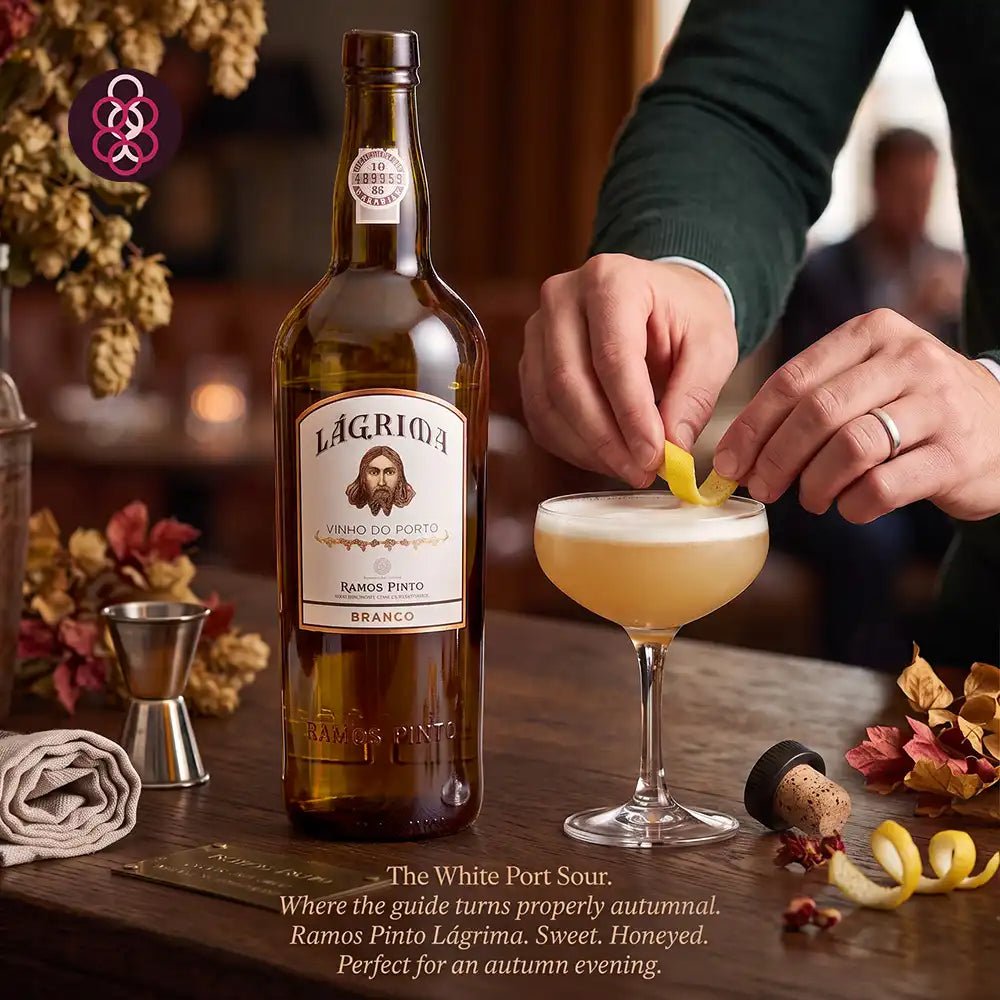 A bottle of Lagrima White Sweet Port cocktail mix stands on a wooden table beside a coupe glass filled with a pale golden drink, while a person's hands garnish the rim with a yellow peel. The scene is warmly lit with autumn-themed decor and a blurred bartender in the background.