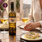 A bottle of Sarmentinho white wine with a colorful label, set on a wooden dining table alongside two wine glasses filled with white wine, plates of food, and a person's hand and arm in a warm, softly lit indoor setting.