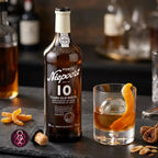 A bottle of Niepoort 10 Years Old White Port wine stands on a dark surface beside a rocks glass filled with amber liquor and an orange peel garnish. Dried orange slices, figs, and a cork are scattered around, with blurred bottles in the background.