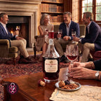 A bottle of Royal Oporto 10 Year Old Tawny Port wine is displayed on a wooden table in a cozy, book-filled living room, surrounded by four people holding wine glasses and enjoying a warm gathering.