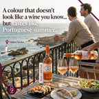 A promotional lifestyle scene on a rocky seaside balcony showing two men looking out at the ocean, with a table in the foreground holding a bottle of rosé wine, two glasses being filled, sushi, grilled fish, and a salad. Large text reads, 'A colour that doesn't look like a wine you know.