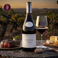 A bottle of Antonio Maçanita FITAPRETA O Iluminista 2024 red wine is displayed on a rustic table outdoors at sunset, alongside a filled wine glass, a cut of raw steak, cheese, and a corkscrew. The background shows a blurred vineyard landscape.
