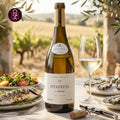 A bottle of white wine from Antonio Maçanita FitaPreta O Ancestral, set on an outdoor table with a wine glass, oysters, a grilled fish, and a salad, with an olive grove in the background.