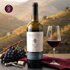 A bottle of Quinta Nova Douro 2024 unaged red wine is displayed on a dark surface next to a filled wine glass, a bunch of grapes, a corkscrew, and a cork, with a blurred vineyard landscape in the background.