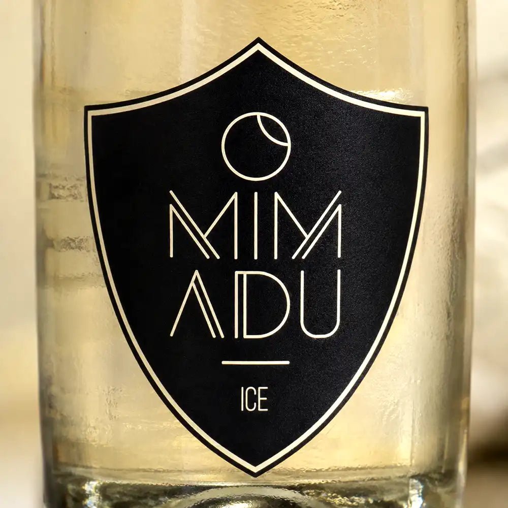 Close-up of a clear glass bottle or tumbler with a black shield-shaped label reading 'MIMADU ICE' in white lettering.
