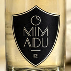 Close-up of a clear glass bottle or tumbler with a black shield-shaped label reading 'MIMADU ICE' in white lettering.