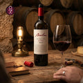 A bottle of Bulas Douro DOC Reserva 2021 red wine stands on a rustic wooden table beside a filled wine glass, with a corkscrew, notebook, and wine barrels in the background. A hand is visible at the lower right.