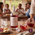 A bottle of Quinta de Vacaria Porto Pink wine is centered on a rustic wooden table, surrounded by glasses of the pink wine, cheese, and cured meats, with a blurred dining scene of people in the background.
