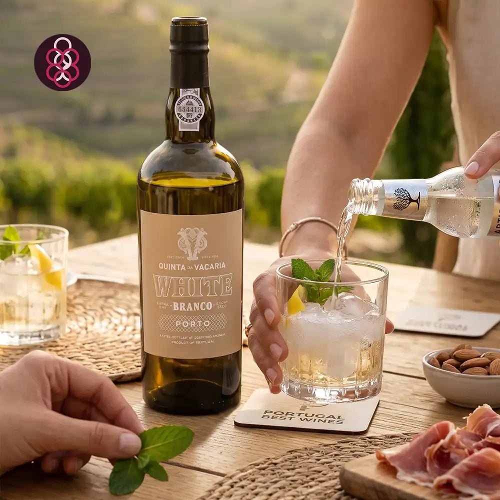 A bottle of Quinta da Vacaria White Branco Porto wine stands on a wooden table beside a glass being filled with the clear liquid from a smaller bottle. Hands are visible holding the glass and garnishing it with mint, with a blurred outdoor vineyard background.