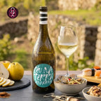 A bottle of Céu na Terra Alvarinho  Vinho Verde White wine with a green label, shown next to a glass of white wine on a dark table with food props, set against a blurred outdoor stone wall background.