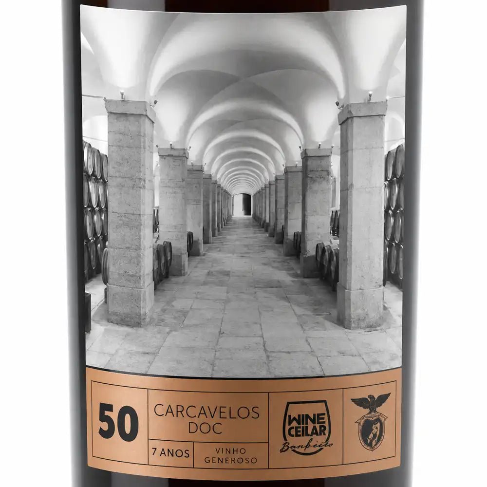 A bottle of wine with a black label featuring a black-and-white photograph of a wine cellar corridor with stone pillars and barrels. The lower label band is tan with black text reading '50 CARCAVELOS DOC', '7 ANOS', and 'VINHO GENEROSO".