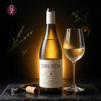 A bottle of Druida white wine with a matching wine glass, a cork, and a corkscrew on a dark stone surface against a dark background with soft botanical accents.
