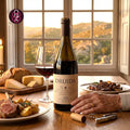 A bottle of Druida red wine stands on a wooden table in a warmly lit dining setting, with a glass of red wine, bread, cheese, and plated food nearby. A person's hand rests near the base of the bottle.