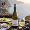 A bottle of FANAL White wine stands on a stone table beside a glass of pale wine, with plates of food and an outdoor coastal landscape in the background. The FANAL logo appears in the upper left corner.