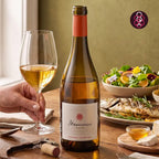 A bottle of Memórias de Santar wine stands on a wooden table next to a hand holding a glass of pale red wine, with a salad, grilled fish, cheese, and crackers in the background.