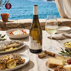 A bottle of white wine stands on a dining table set with a glass of white wine, plates of grilled fish and pasta, cheese, and an outdoor seaside background.