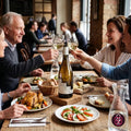 A group of people dining together in a restaurant or cafe setting, clinking wine glasses around a bottle of white wine on a wooden table with plates of food.