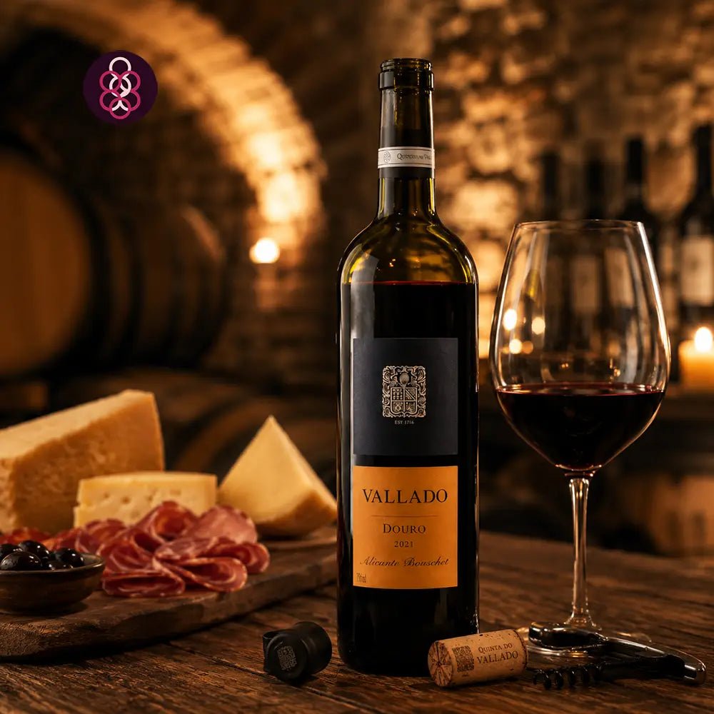 A bottle of Vallado Alicante Bouschet red wine stands on a wooden table beside a filled wine glass, cheese wedges, sliced salami, and a corkscrew, with a blurred wine cellar background and a purple logo in the top left corner.