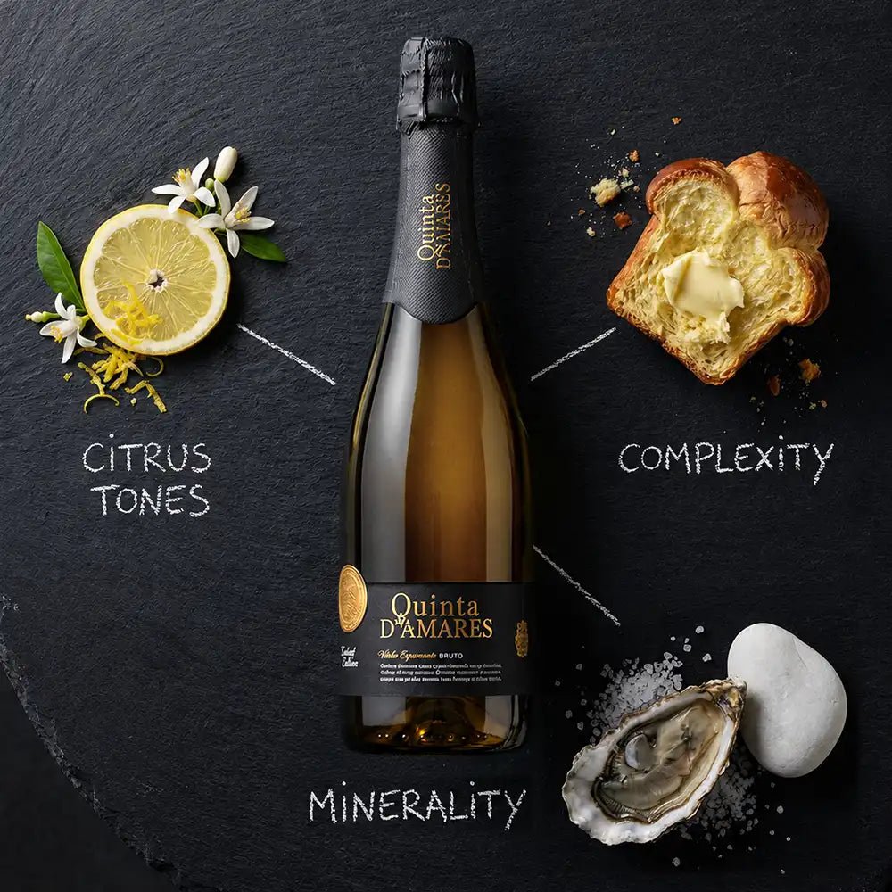 A bottle of Quinta D'Amares sparkling wine centered on a dark slate background, surrounded by styled food and ingredient props: a lemon slice with white flowers labeled 'Citrus Tones,' a slice of bread with butter labeled 'Complexity,' and an oyster with salt and stone labeled 'Minerality.' Visible brand text on the bottle and label reads Quinta D'Amares.