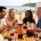 Four people laughing and drinking from a bottle of Lancers Sangria on a beach table, with a beach and ocean in the background.