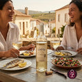 A bottle of Canto Décimo white wine is set on a rustic wooden table between two smiling women holding wine glasses, with plates of grilled fish and salad in the foreground and a vineyard landscape in the background.