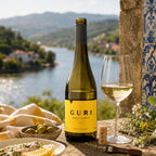 A bottle of Guri white wine with a yellow label stands on a stone ledge beside a filled wine glass, olives, lemon slices, and a corkscrew, with a scenic river and mountains in the background.
