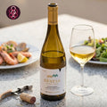 A bottle of Génese Alvarinho white wine stands on a speckled countertop beside a filled wine glass, with plates of food blurred in the background. A corkscrew and cork are in the foreground.