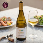 A bottle of Génese Alvarinho white wine stands on a speckled countertop beside a filled wine glass, with plates of food blurred in the background. A corkscrew and cork are in the foreground.