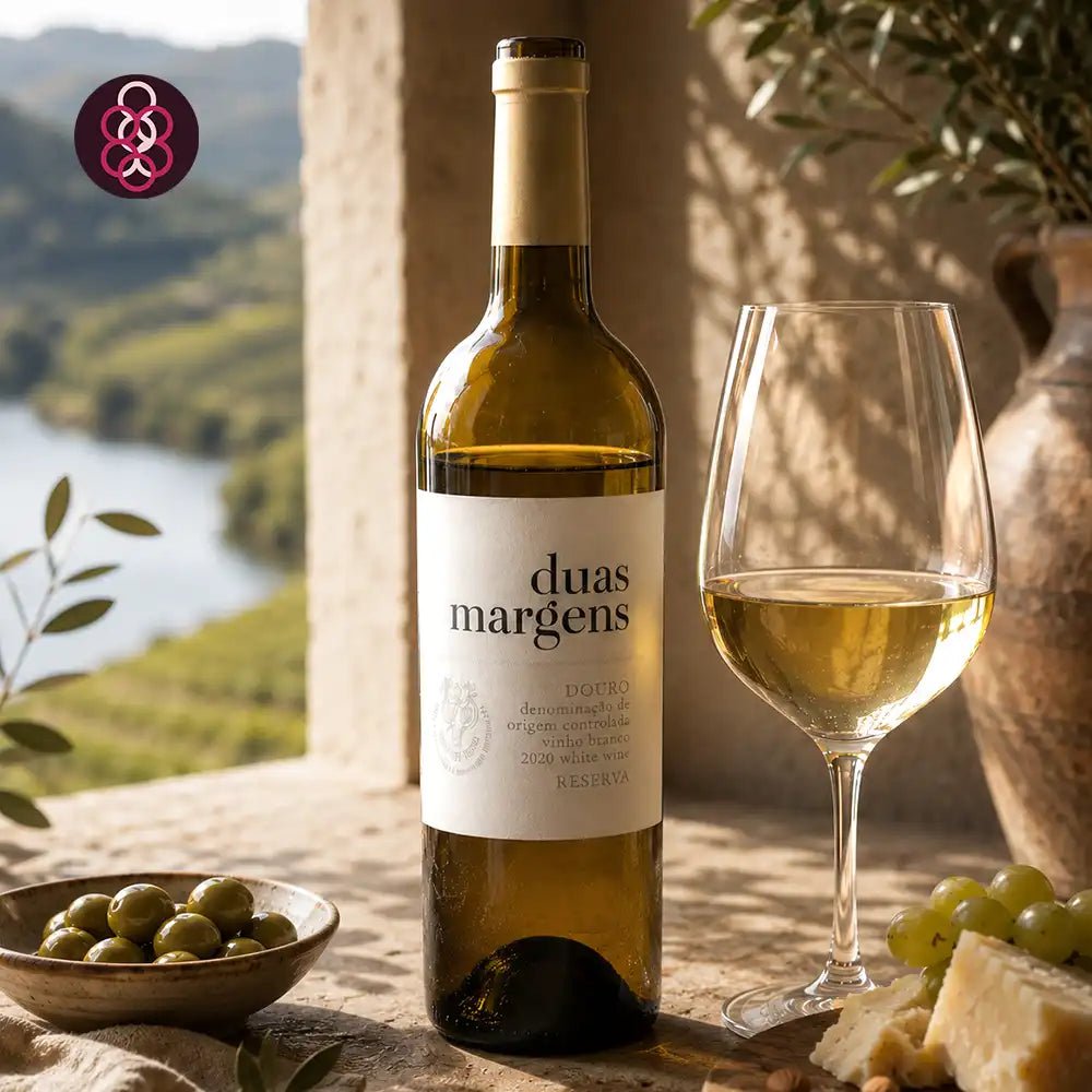 A bottle of duas margens white wine stands on a stone surface beside a filled wine glass, with olives, cheese, grapes, and an outdoor landscape visible in the background.