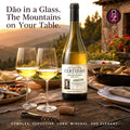 A bottle of white wine with two filled wine glasses on an outdoor dining table at sunset, with food dishes and a stone building in the background. Portuguese marketing text appears around the product.