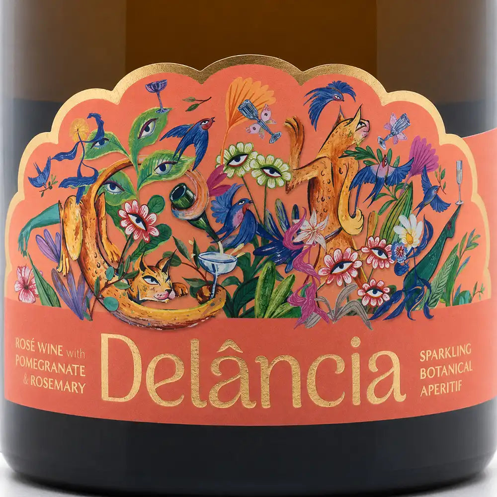 Close-up of a Delância sparkling apéritif wine bottle label, featuring a vibrant, whimsical botanical illustration with a cat, birds, and flowers on a peach-orange background with gold foil lettering.