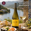 A bottle of Guri white wine with a yellow label stands on a stone ledge beside a filled wine glass, olives, lemon slices, and a corkscrew, with a scenic river and mountains in the background.