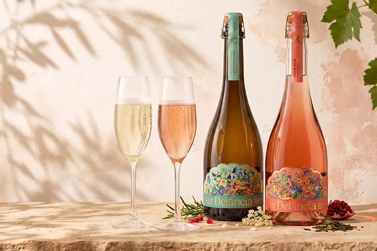 Two Delancia sparkling wine bottles and two filled champagne flutes on a rustic stone surface against a warm beige wall with leaf shadows.