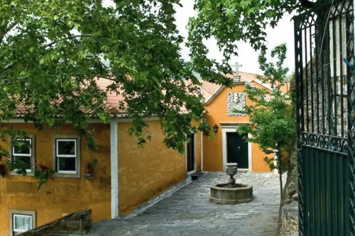 Quinta_do_Vallado-A scenic view of a historic yellow-orange building with a terracotta tiled roof, featuring a stone courtyard with a small fountain, lush green trees, and a dark wrought-iron gate on the right.