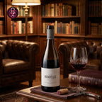 A bottle of Manoella Douro red wine stands on a wooden table in a cozy library setting, alongside a filled wine glass, a cork, and a corkscrew. The background features warm leather armchairs and wooden bookshelves.